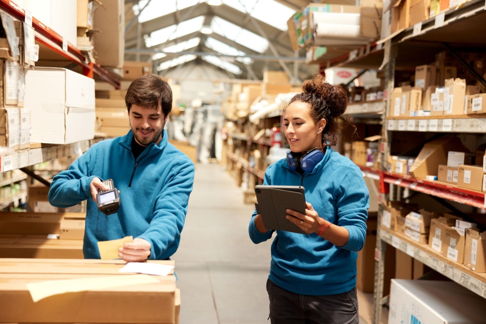 Logistique commerce : deux opérateurs en entrepôt, l’un scanne un colis, l’autre utilise une tablette, avec des rayonnages remplis de cartons en arrière-plan.