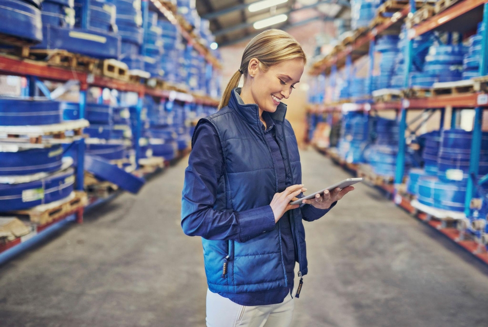 Mujer en un almacén utilizando una tablet, rodeada de estanterías con grandes bobinas de cables azules y equipos industriales, gestionando una orden de transporte digital