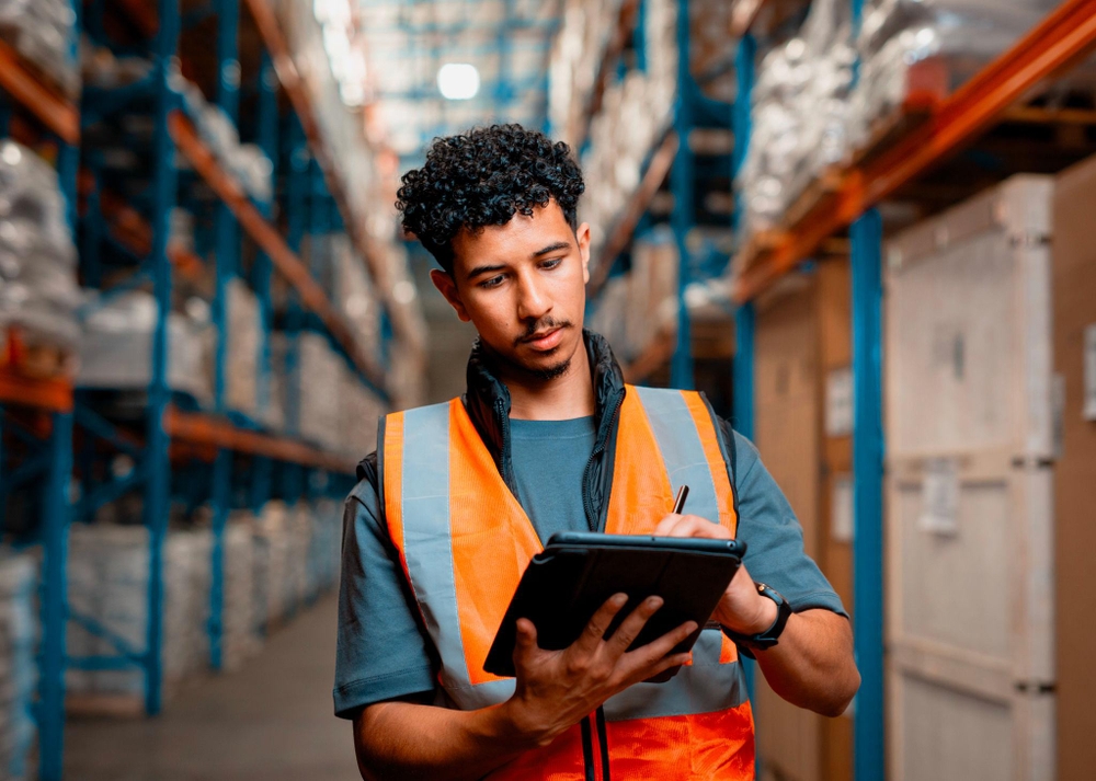 Un opérateur en gilet de sécurité orange utilise une tablette dans un entrepôt aux rayonnages élevés remplis de marchandises, illustrant la logistique commerce.