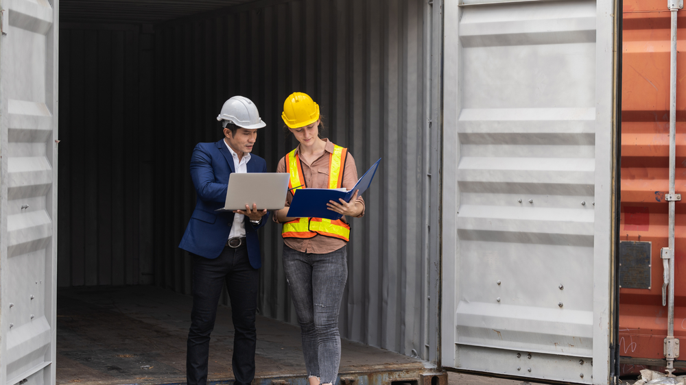 Deux personnes en casque de chantier examinant une facture transport sur ordinateur portable à l’intérieur d’un conteneur d’expédition