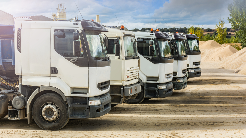 Cinco camiones volquete blancos estacionados en fila en una obra con montones de arena y árboles al fondo, representando la actividad regulada por la LOTT en el transporte terrestre.