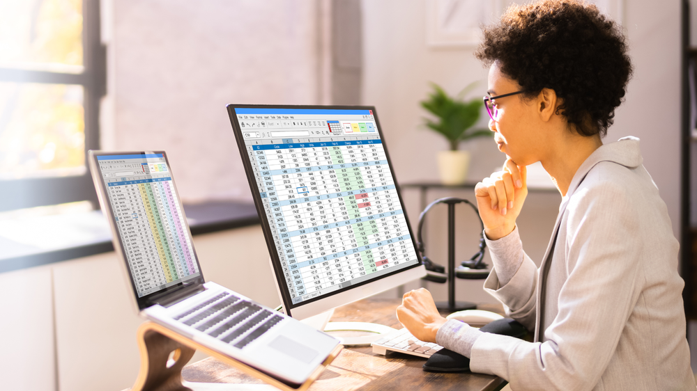 Analyste vérifiant une facture transport sur deux écrans avec tableur et ordinateur portable dans un bureau lumineux