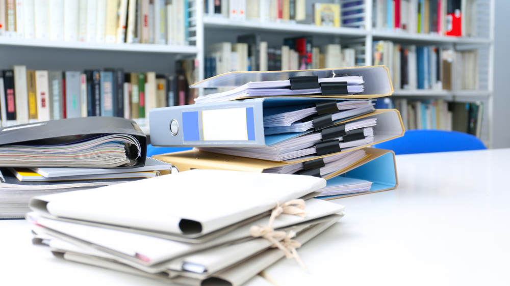 Pilas de archivadores y documentos sobre una mesa blanca en una biblioteca, con estanterías llenas de libros al fondo, simbolizando la gestión administrativa y documental que regula la LOTT.