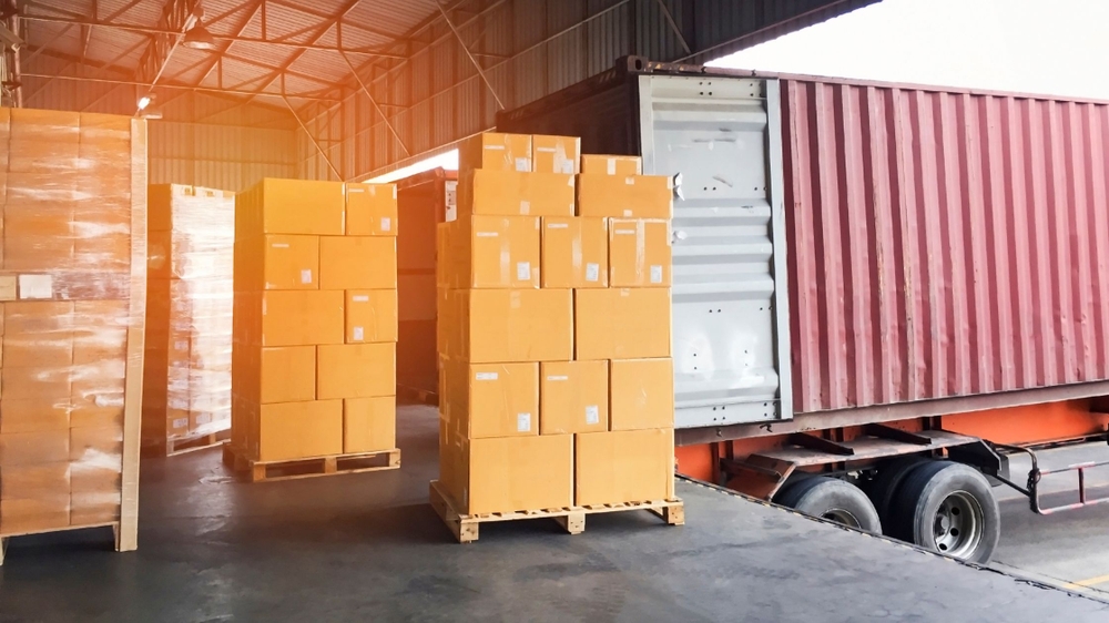 Palets con cajas de cartón en un almacén, representando un proceso de logística de distribución con un camión cargando un contenedor al fondo.