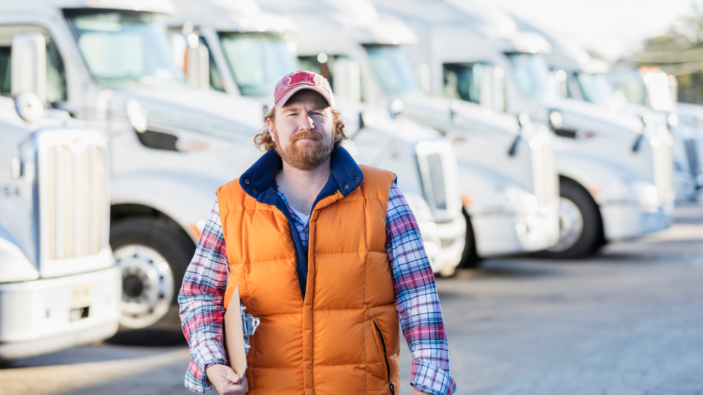gestion de flotte poids lourds un homme en gilet orange et casquette rouge tient un clipboard devant une rangée de camions blancs