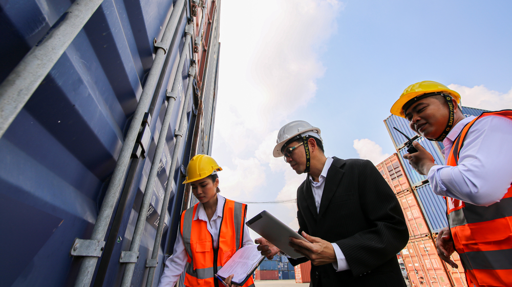 Trois travailleurs en tenue de sécurité inspectent un conteneur, avec clipboard, documents et radio, illustrant la coordination logistique avec WMS TMS.