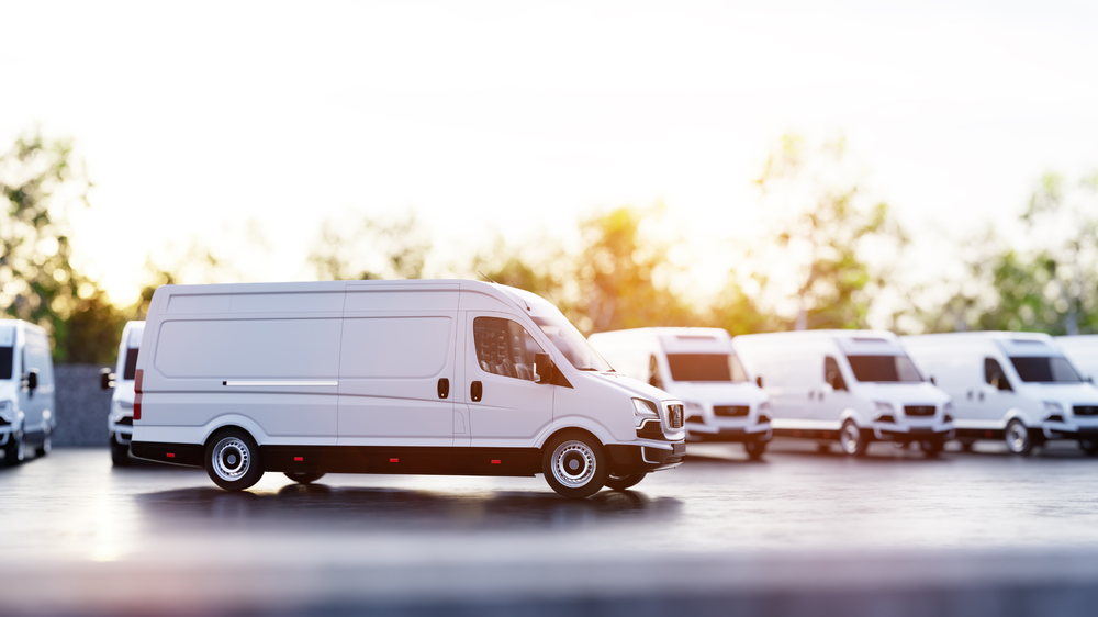 Fila de furgonetas blancas de reparto estacionadas en un aparcamiento con árboles al fondo, iluminadas por una luz suave, representando la flota regulada por la LOTT.