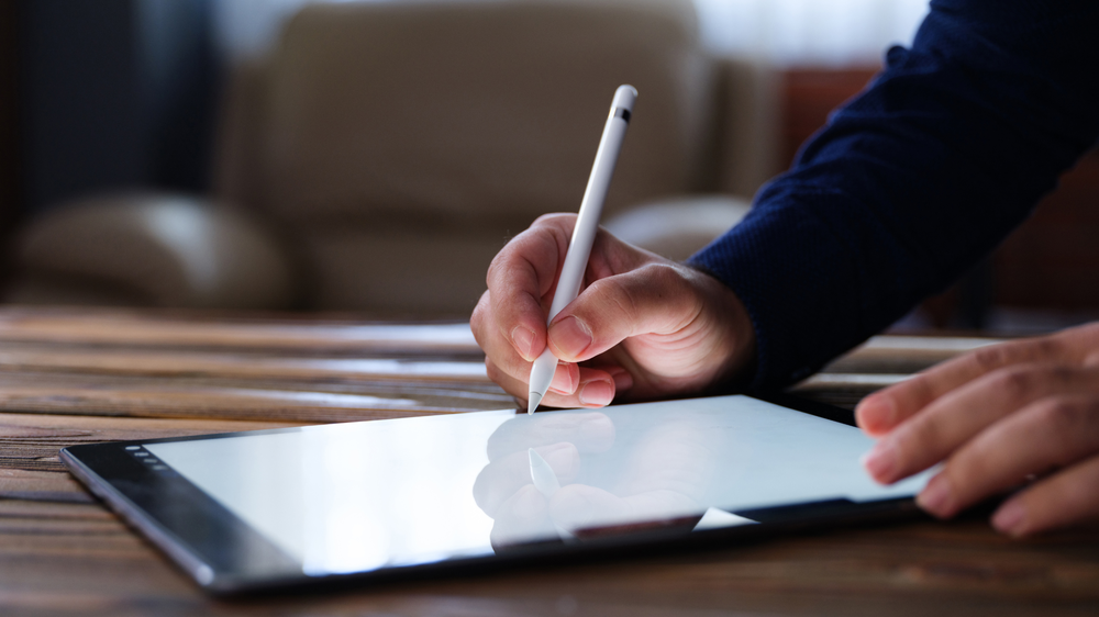 Persona utilizando un lápiz digital para escribir en la pantalla de una tableta sobre una mesa de madera, símbolo de la digitalización documental promovida por la LOTT.