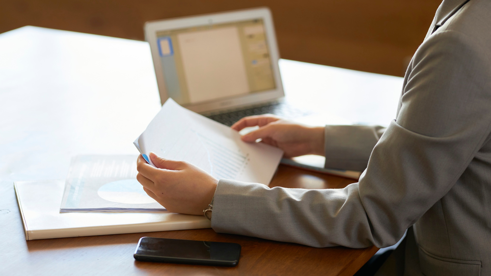 Persona con traje revisa documentos en un escritorio con un portátil y un smartphone, simbolizando la gestión documental de los tipos de aduanas.