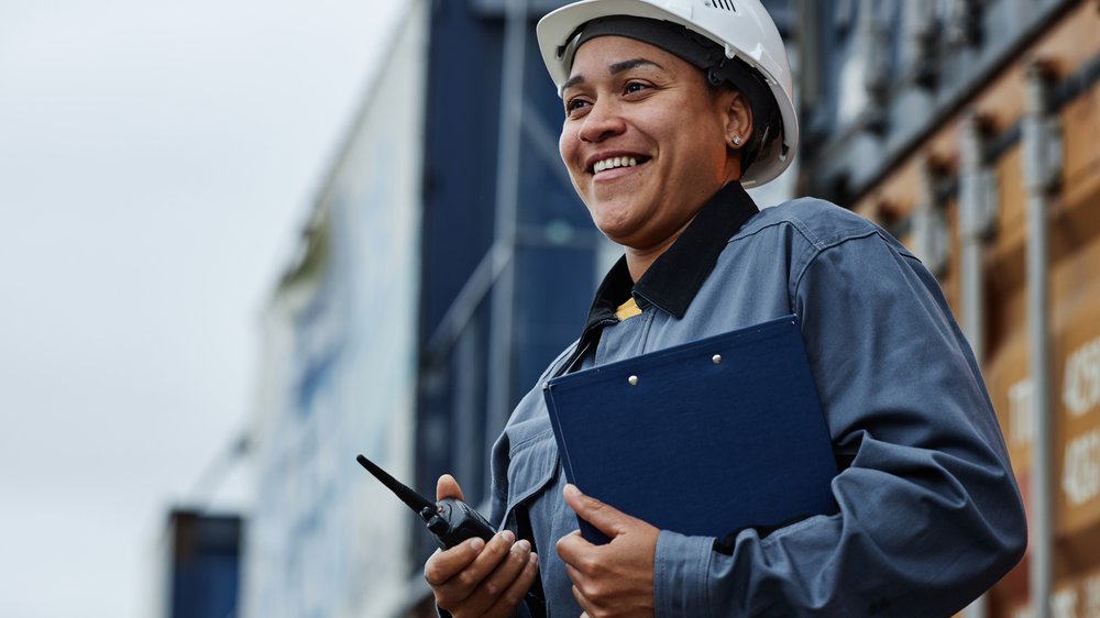 Trabajador sonriente con casco, portapapeles y walkie-talkie, de pie junto a contenedores de carga, representando la coordinación en la externalización logística.