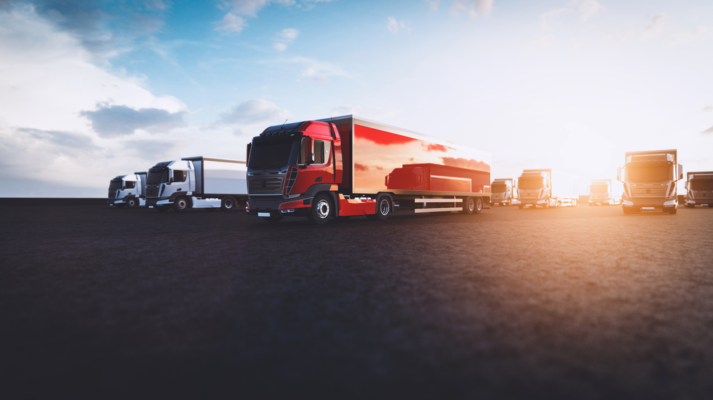 gestion de flotte poids lourds rangée de camions stationnés sur une grande surface sombre sous un ciel bleu avec des nuages dispersés et le soleil brillant à l’horizon