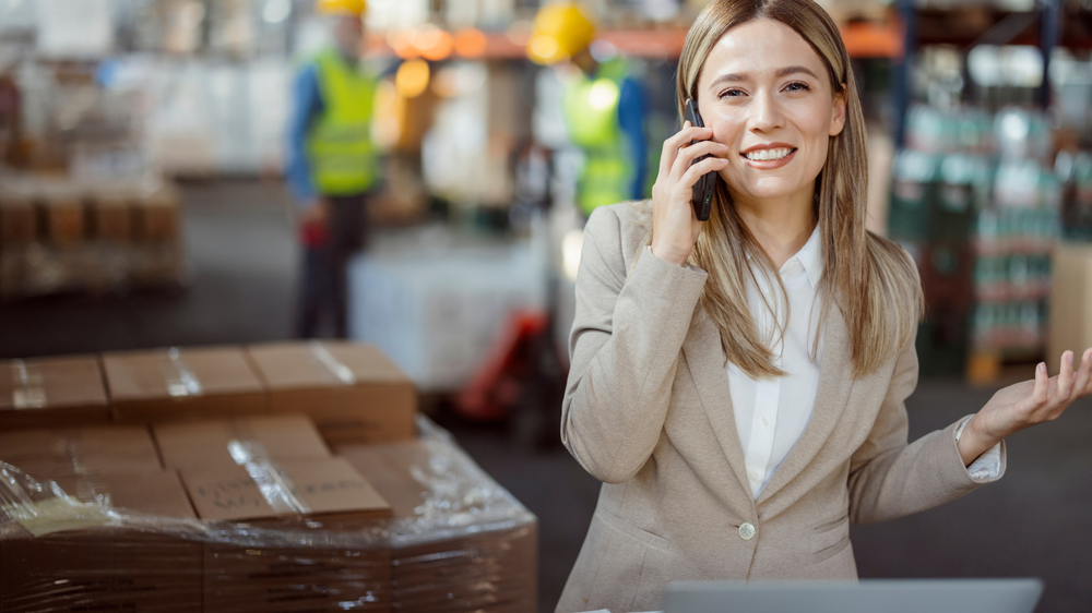 Femme souriante dans un entrepôt, au téléphone, au cœur d’une opération d’externalisation logistique, entourée de palettes empilées et de travailleurs en gilet de sécurité en arrière-plan.