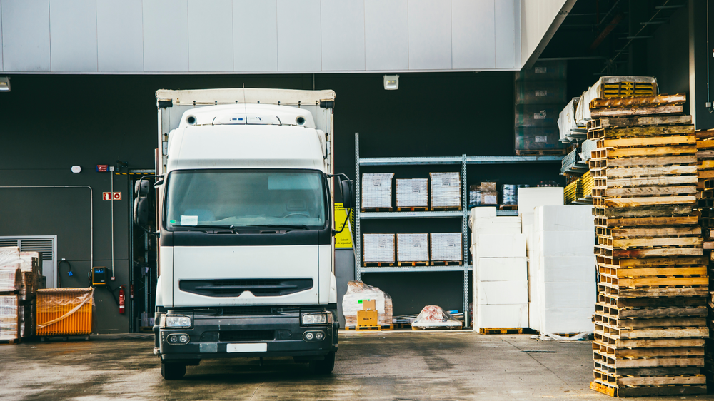 Un camion blanc stationné à un quai de chargement, avec des palettes empilées et des étagères de cartons, illustrant l’importance de la liste de colisage dans la gestion logistique.