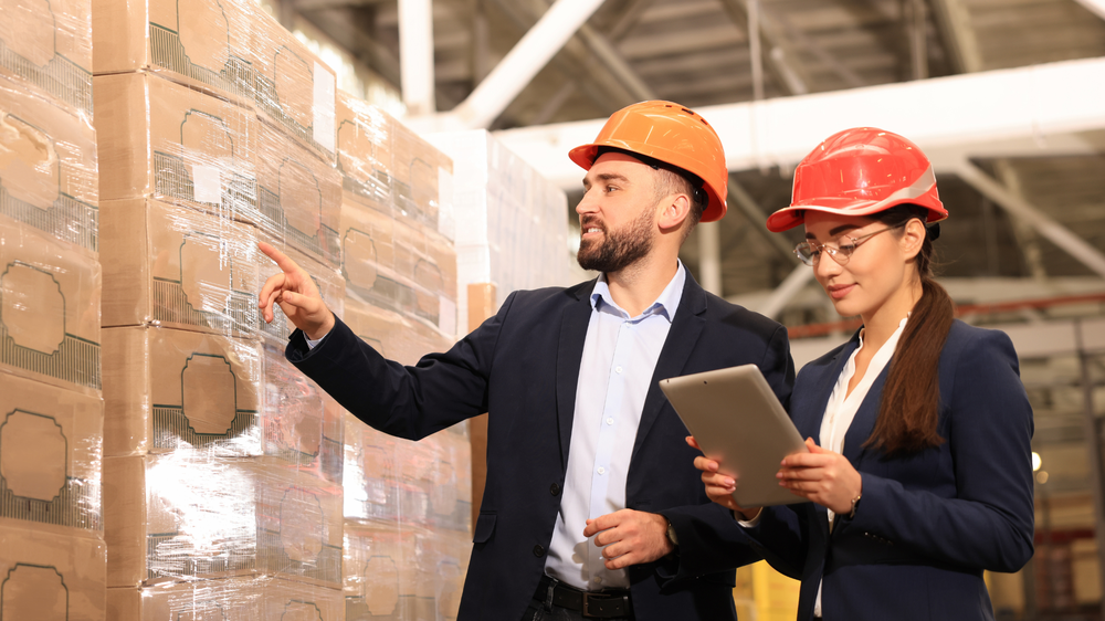 Deux personnes en casque et en costume dans un entrepôt ; l’une pointe du doigt pendant que l’autre tient une tablette, debout près de palettes empilées — externalisation logistique.