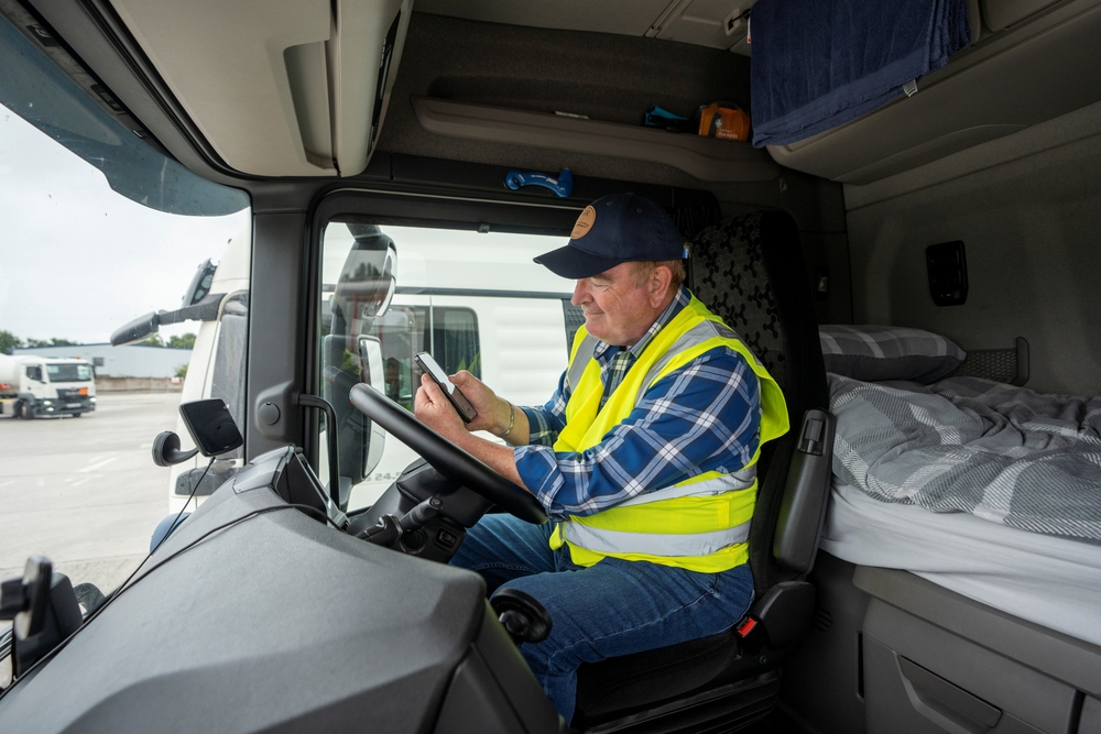 Un conductor de camión con chaleco reflectante está sentado en la cabina, revisando su teléfono, en un espacio equipado con cama y objetos esenciales, ilustrando el día a día en la gestión de flotas de transporte.