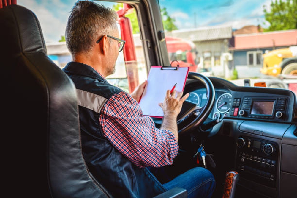 Homme assis dans le siège conducteur d’un camion, portant une chemise à carreaux et un gilet, écrivant sur une lettre de voiture électronique sur un clipboard. Tableau de bord et volant visibles.