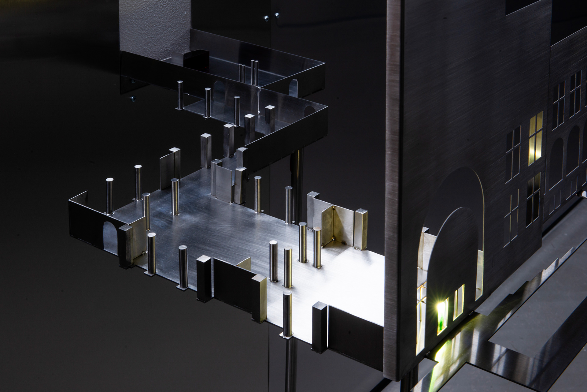 Metallic architectural model with a linear design, featuring columns, arches, and illuminated windows against a dark background.