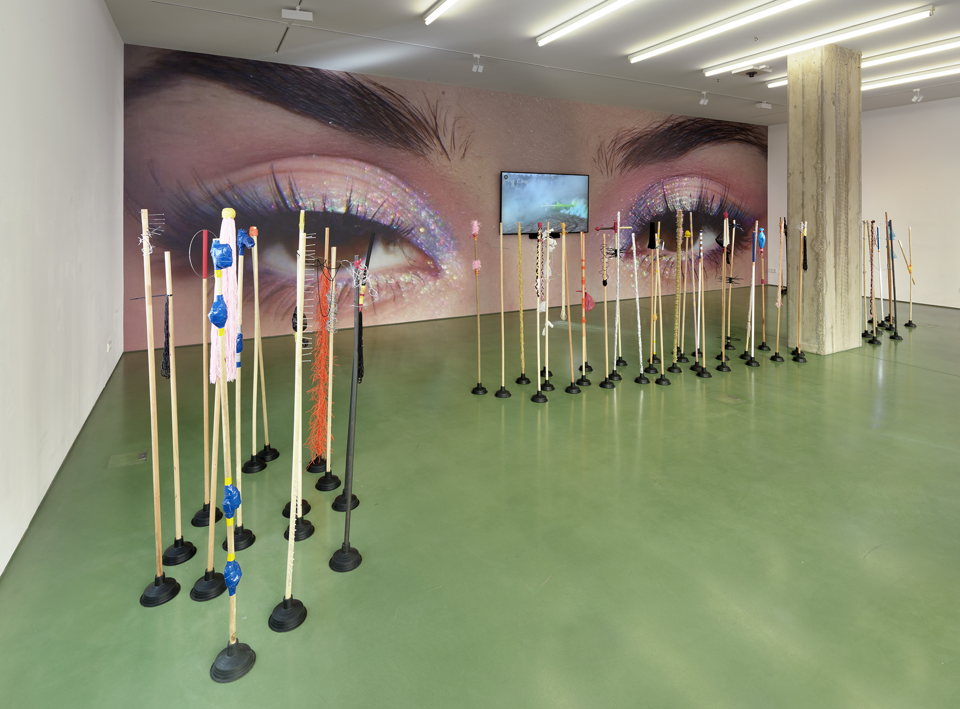 Art installation with decorated poles on a green floor, large mural of glittery eyes in the background, and a wall-mounted screen.