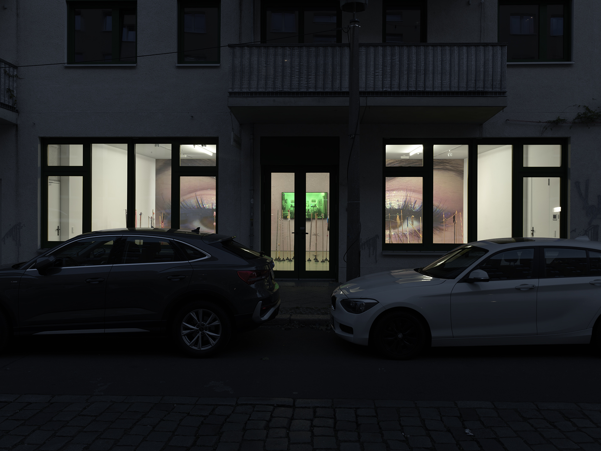 A dimly lit street with two parked cars in front of a building displaying illuminated artwork through large windows.