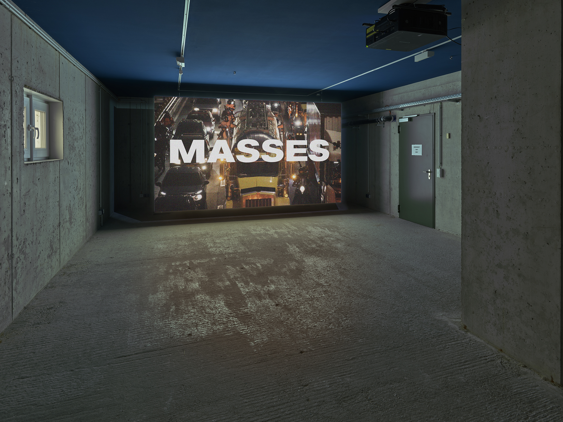 A dimly lit concrete room with a large screen displaying the word "MASSES" over a busy highway scene.