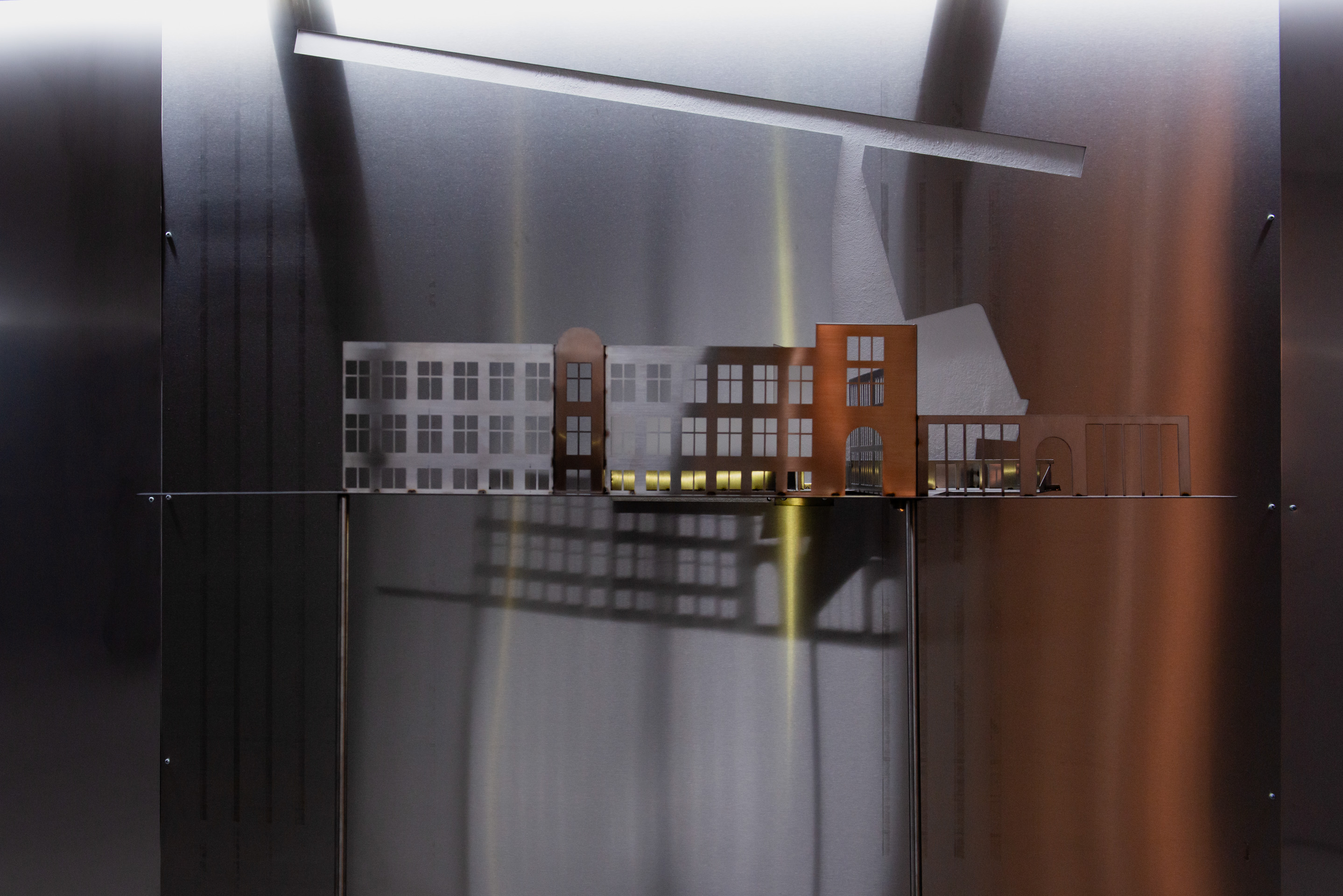 A metal architectural model with cut-out windows casts shadows on a reflective surface, illuminated by a soft overhead light.