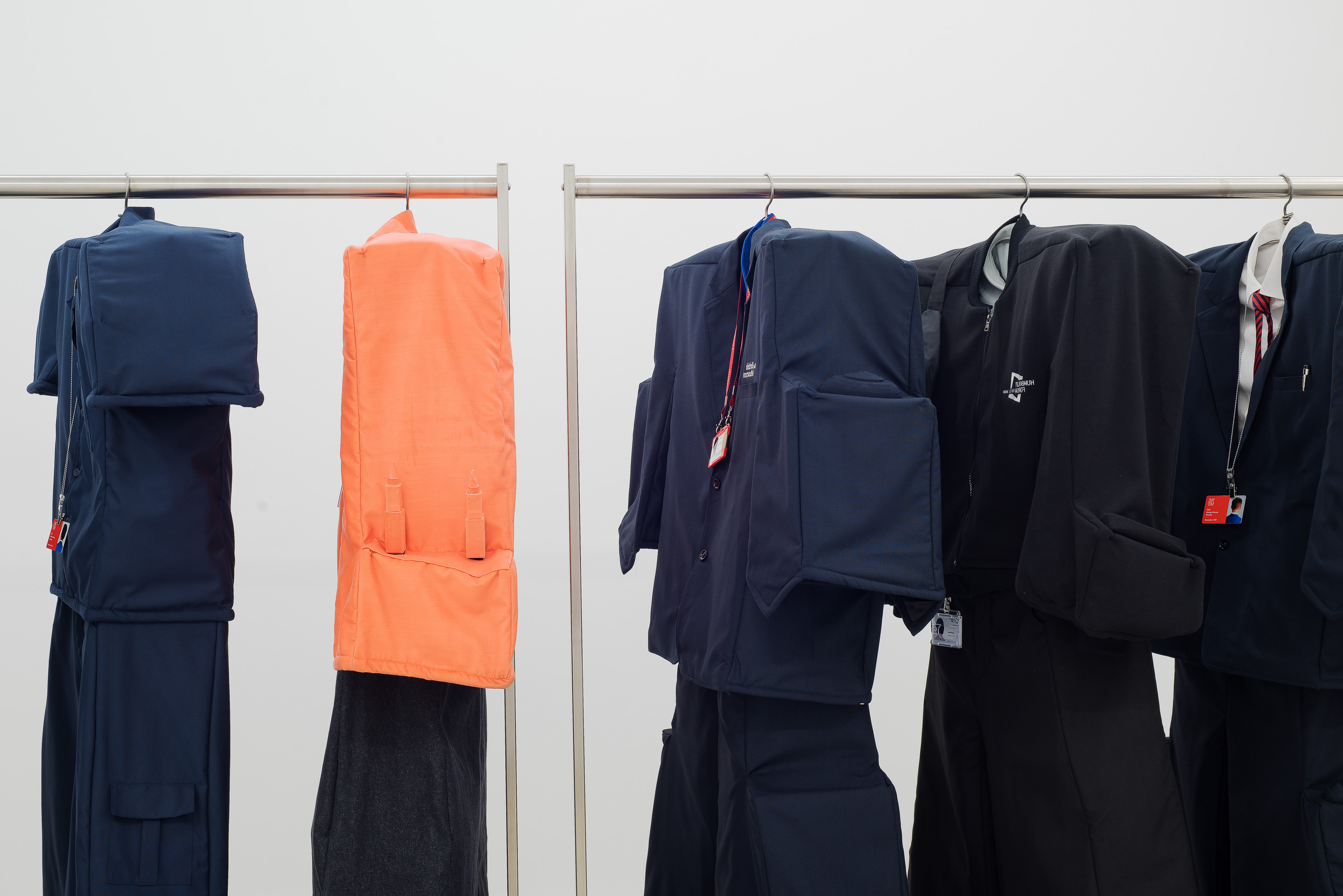 Several sets of padded clothing, including blue and orange, hang on a metal rack. They have attached ID badges and tags.