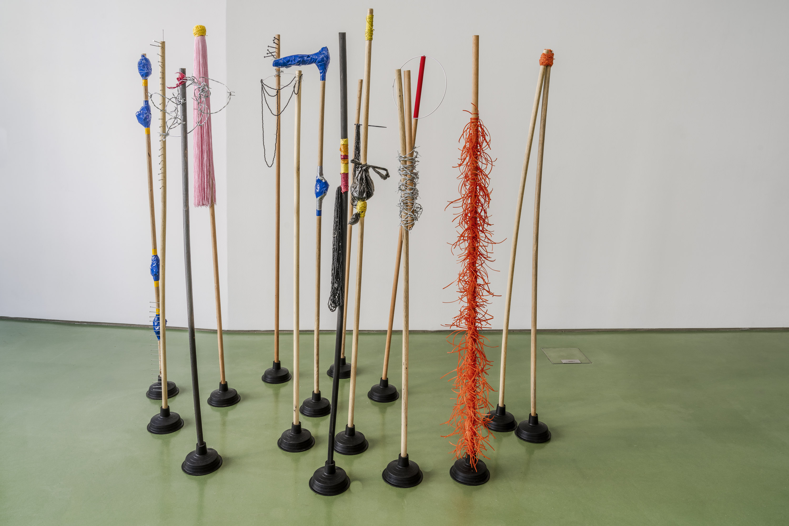 A group of sculptures with tall wooden handles, each adorned with colorful materials like yarn, wire, and feathers, arranged on a green floor.