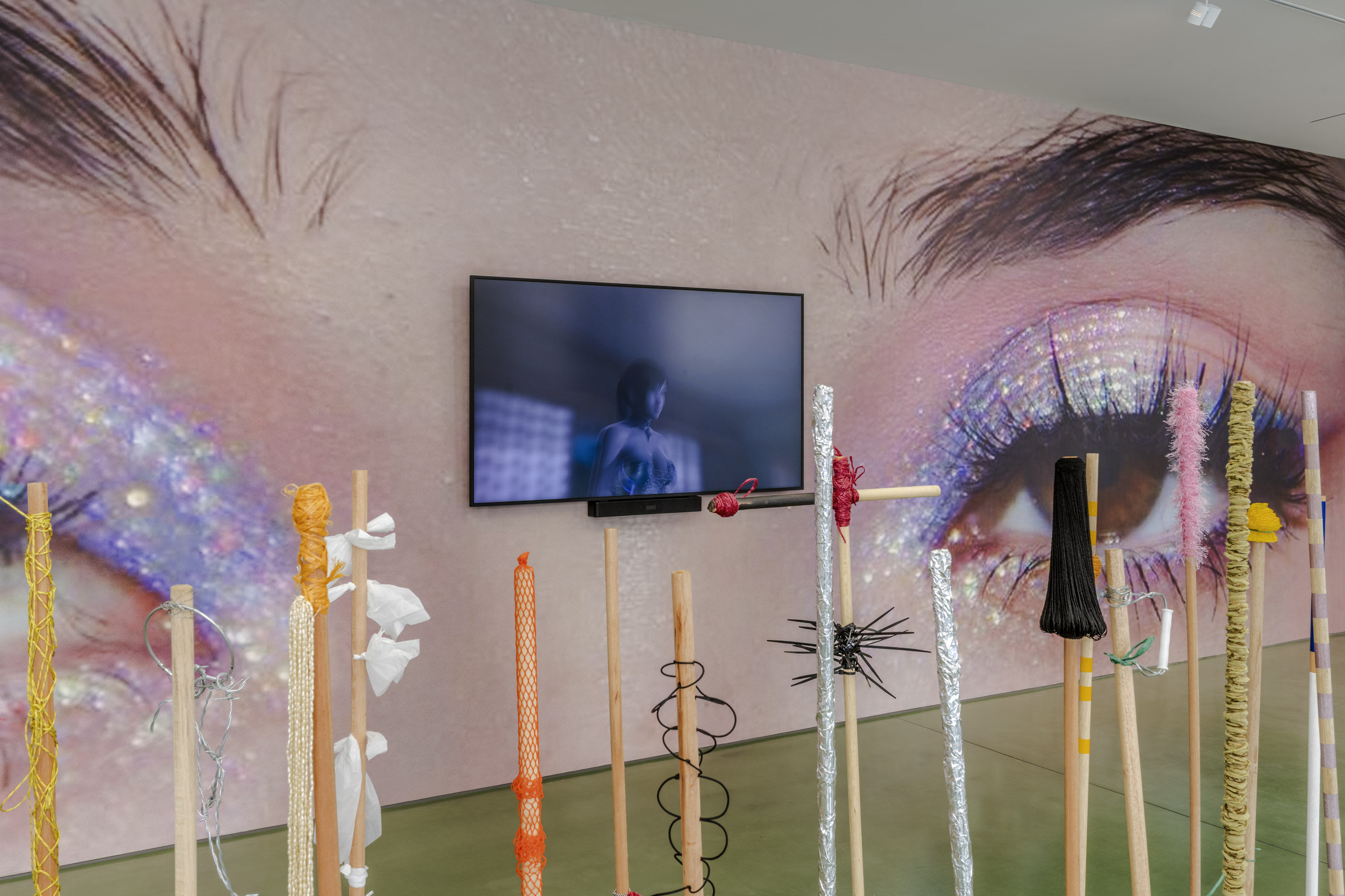 Art installation with colorful poles in the foreground and a large screen showing a close-up of glittery eye makeup in the background.
