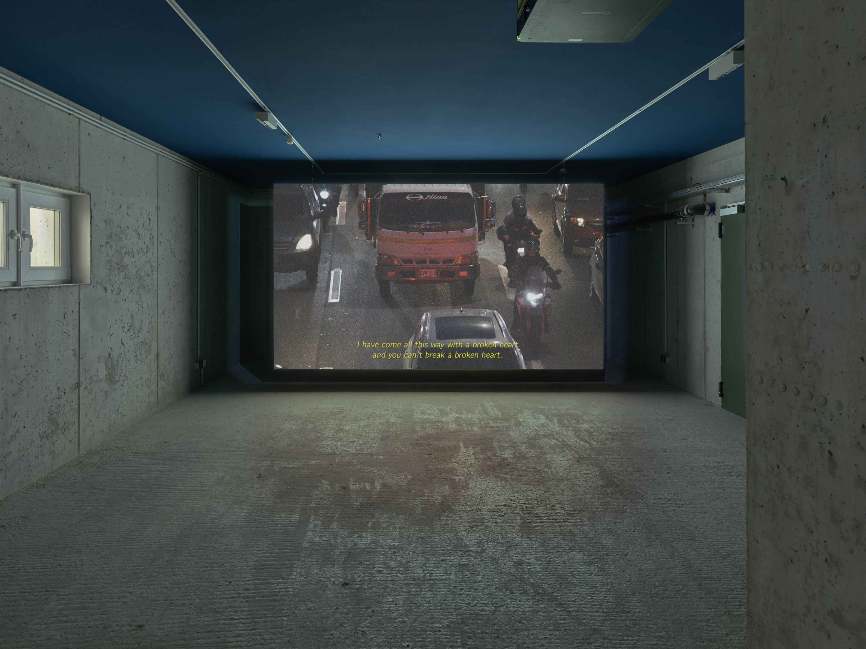 A dimly lit room with a large screen displaying a street scene featuring vehicles. Subtitles read: "I have come to give you my love and you didn't break a broken heart."
