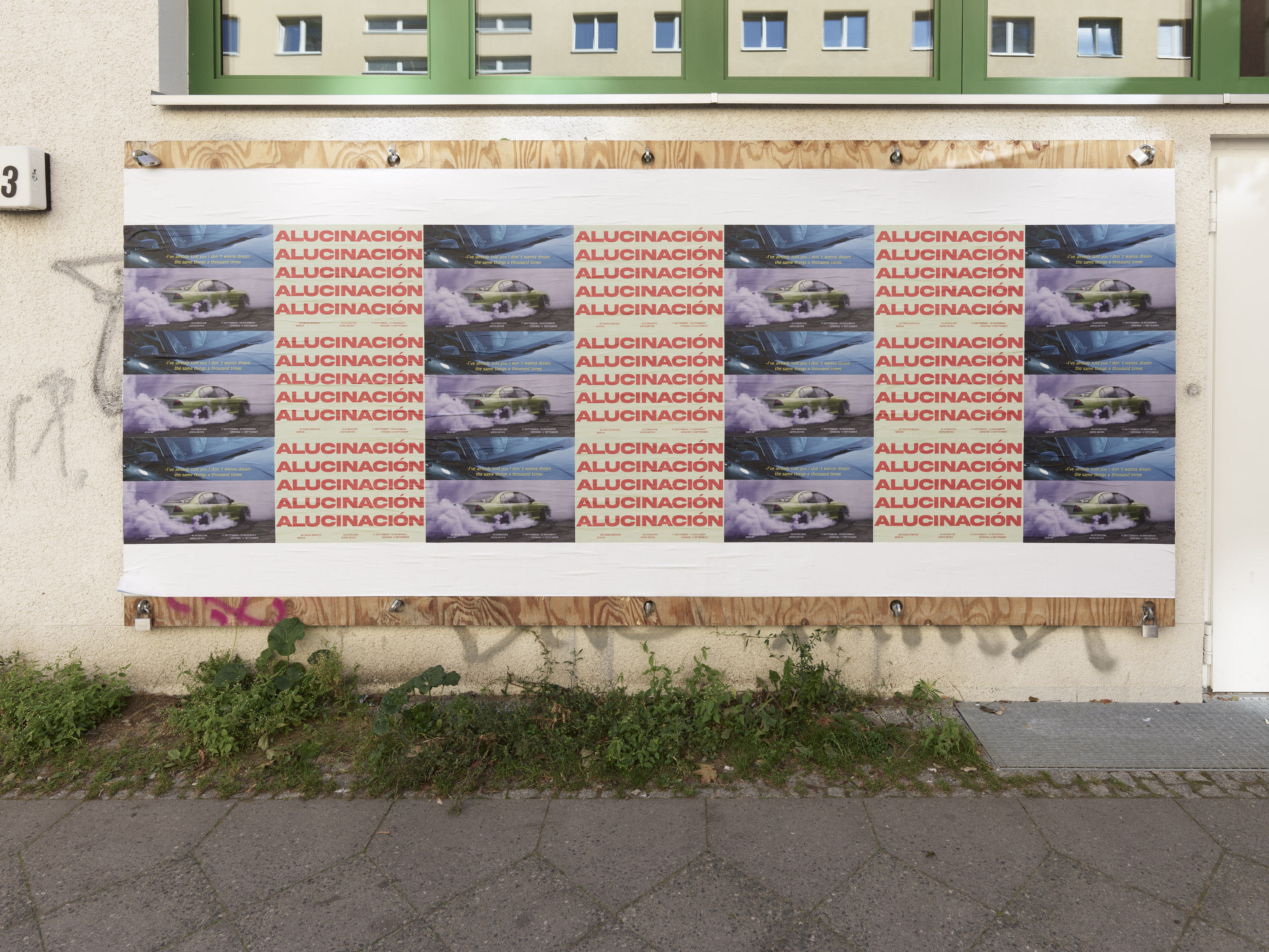 A wall display with repeating posters featuring the word "ALUCINACION" and images of a speeding car. Green windows are above the posters.
