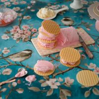 Waffle ice cream sandwiches with pink filling on a floral tablecloth featuring birds and blossoms, surrounded by plates and a spoon.