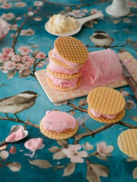 Waffle ice cream sandwiches with pink filling on a floral tablecloth featuring birds and blossoms, surrounded by plates and a spoon.