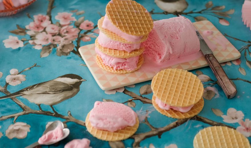 Waffle ice cream sandwiches with pink filling on a floral tablecloth featuring birds and blossoms, surrounded by plates and a spoon.