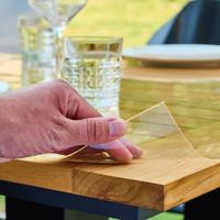 A hand peeling protective film from a wooden table, with glasses and plates set on top.
