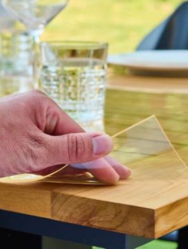 A hand peeling protective film from a wooden table, with glasses and plates set on top.