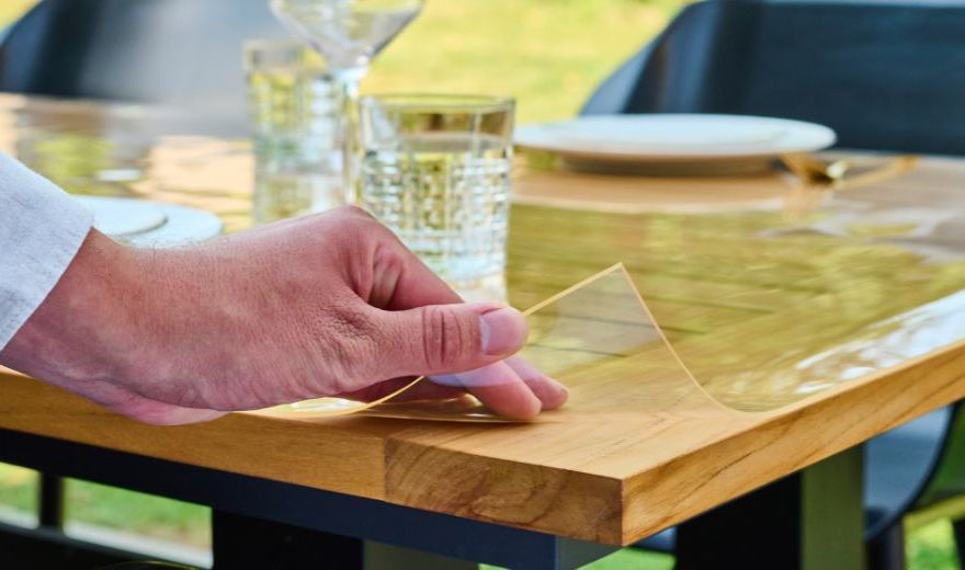 A hand peeling protective film from a wooden table, with glasses and plates set on top.