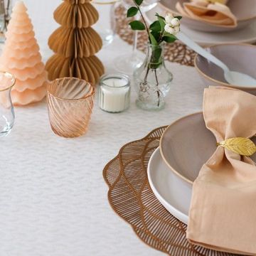 Elegant table setting with beige napkins, leaf-shaped napkin rings, glassware, candles, and decorative trees on a textured tablecloth.