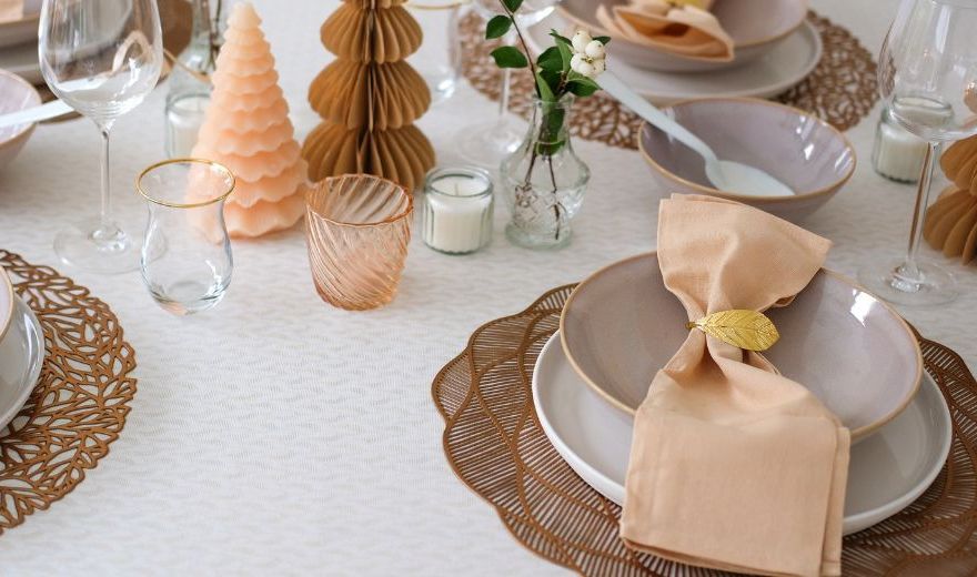 Elegant table setting with beige napkins, leaf-shaped napkin rings, glassware, candles, and decorative trees on a textured tablecloth.