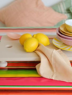 A colorful table with a striped cloth, stacked bowls and plates, lemons, a beige napkin, and a yellow jug. Cushions in the background.