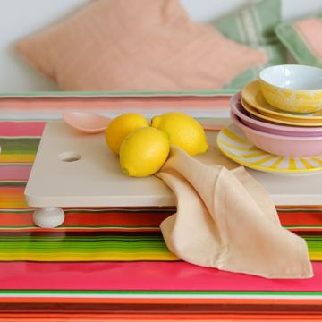 A colorful table with a striped cloth, stacked bowls and plates, lemons, a beige napkin, and a yellow jug. Cushions in the background.