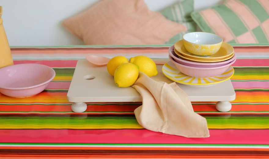 A colorful table with a striped cloth, stacked bowls and plates, lemons, a beige napkin, and a yellow jug. Cushions in the background.