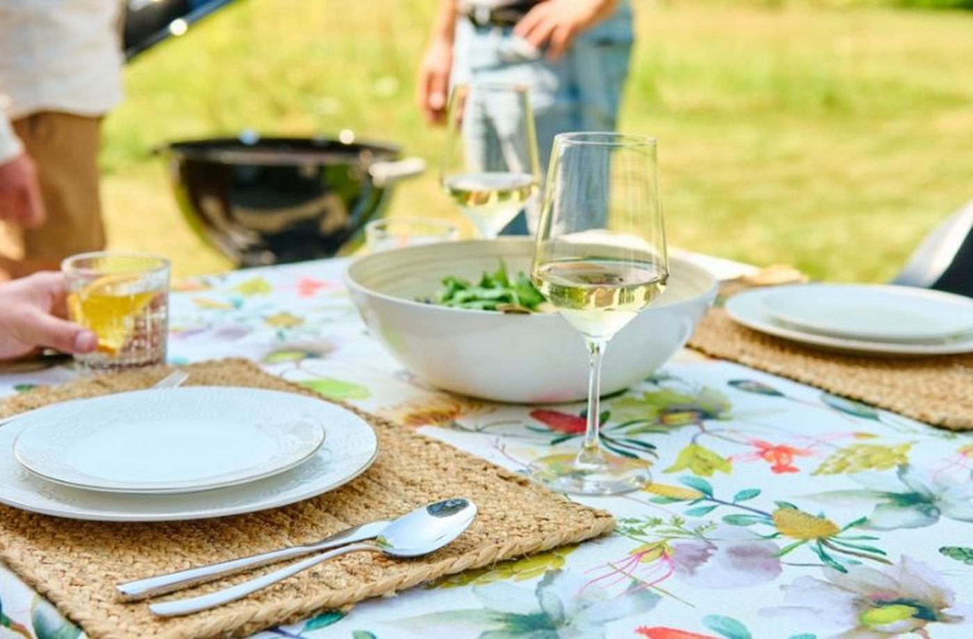 Outdoor table with floral cloth set for two, featuring plates, cutlery, a salad bowl, and wine glasses; people and a grill in the background.