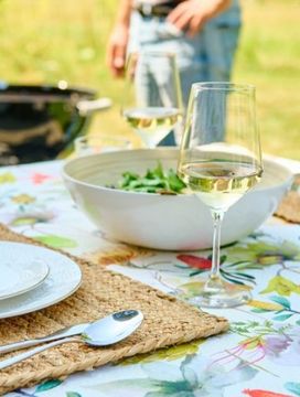 Outdoor table with floral cloth set for two, featuring plates, cutlery, a salad bowl, and wine glasses; people and a grill in the background.