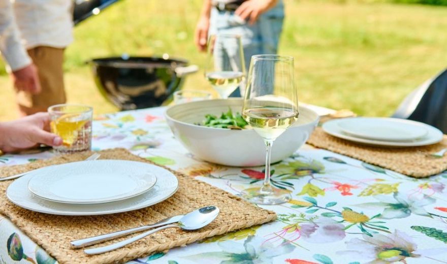 Outdoor table with floral cloth set for two, featuring plates, cutlery, a salad bowl, and wine glasses; people and a grill in the background.