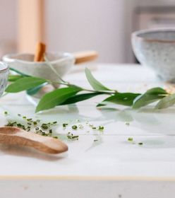 Ceramic bowls and small dishes with herbs and cinnamon sticks on a white table, accented by green leaves.