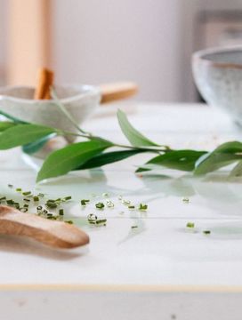 Ceramic bowls and small dishes with herbs and cinnamon sticks on a white table, accented by green leaves.