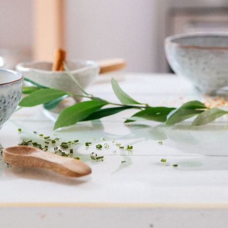 Ceramic bowls and small dishes with herbs and cinnamon sticks on a white table, accented by green leaves.