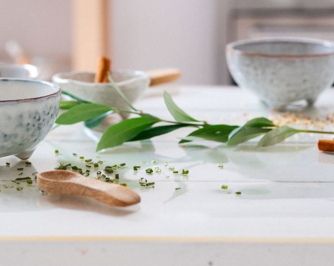 Ceramic bowls and small dishes with herbs and cinnamon sticks on a white table, accented by green leaves.