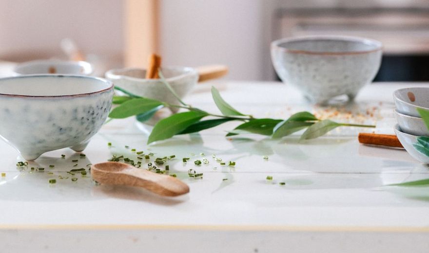 Ceramic bowls and small dishes with herbs and cinnamon sticks on a white table, accented by green leaves.