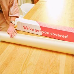 Person unrolling white wrapping paper from a red box labeled "We’ve got you covered" on a wooden table.